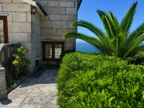 Entrada de villa de piedra en Oia con palmera, jardín exuberante y vista al mar en día despejado