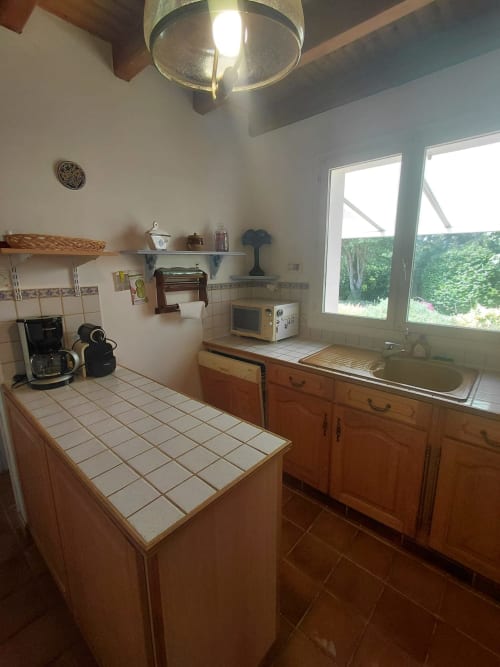 Cuisine lumineuse de cottage à Locmaria avec meubles en bois, comptoirs carrelés, évier, micro-ondes et vue sur le jardin