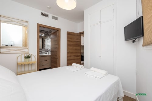 Modern bedroom at Apartamentos Manbea in Almería with white bedding, wooden doors, wall-mounted TV, and minimalist décor