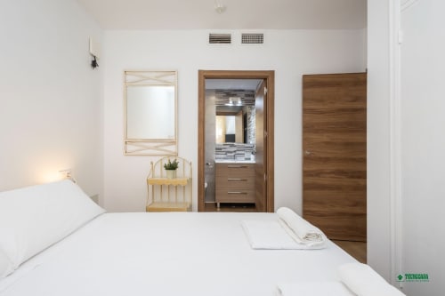 Bright bedroom at Apartamentos Manbea in Almería with white bedding, wooden bathroom door, and mirror.