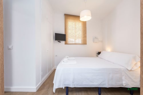 Manbea 11 bedroom in Almería with white bedding, wall-mounted TV, ceiling lamp, and wooden window blinds.