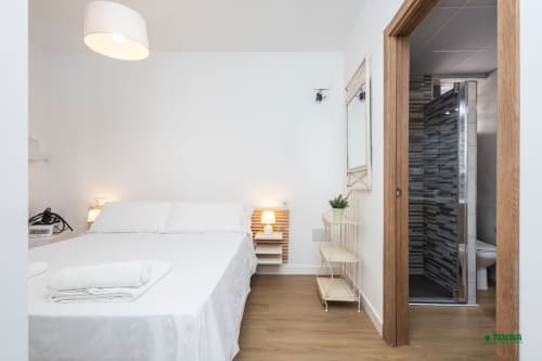 Modern bedroom at Apartamentos Manbea in Almería with white bed, wooden nightstand, and ensuite bathroom with dark tile