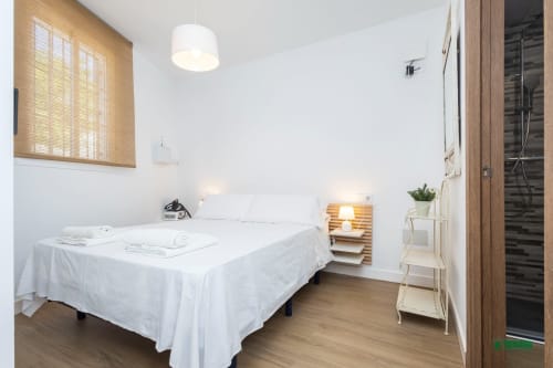 Bright bedroom at Apartamentos Manbea in Almería with white bedding, wooden nightstand, pendant light, and modern bathroom