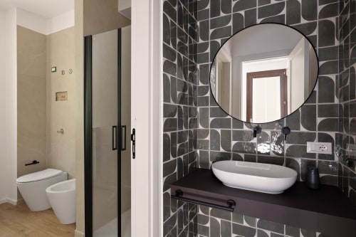 Bagno moderno a Dimora Silva Nature Suites con rivestimento geometrico nero, specchio rotondo, lavabo bianco, toilette e vetro