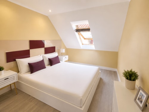 Acogedor dormitorio abuhardillado en el hotel Holibai de Baiona con paredes crema, cabecero burdeos, claraboya y suelos de madera.