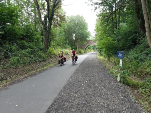Cyclists on tree-lined canal path near Villa Bianca in Dalhem, Belgium with brick bridge ahead