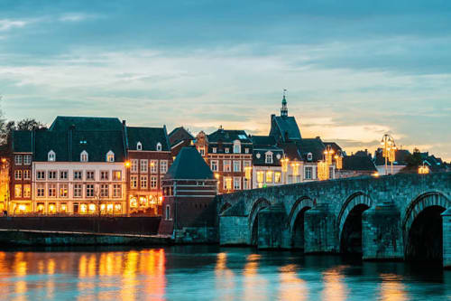 Historic European riverfront with illuminated buildings and stone bridge at dusk in Maastricht area