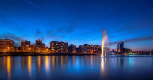 Evening cityscape with illuminated buildings reflecting in calm water and fountain, near Villa Bianca in Mortroux Dalhem