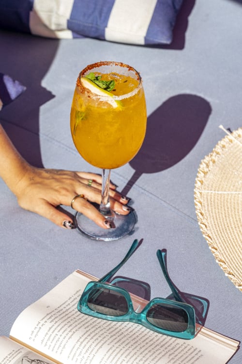 Cóctel dorado con borde especiado y garnish de lima en Hotel Isla Mujeres Centro, sostenido sobre libro abierto y gafas de sol