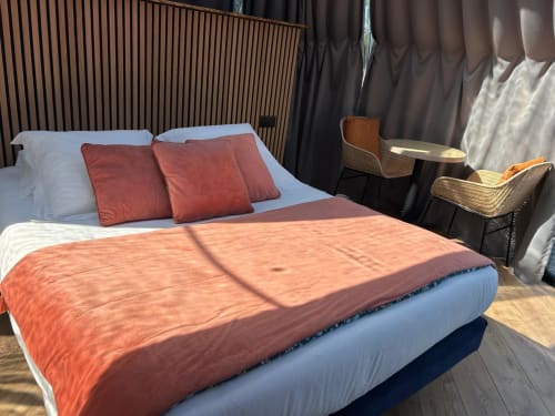 Chambre Bulle Topaze à l'hôtel Bubble Jade avec couverture orange, oreillers rouges, tête de lit en bois et vue sur le jardin à Bazouges la