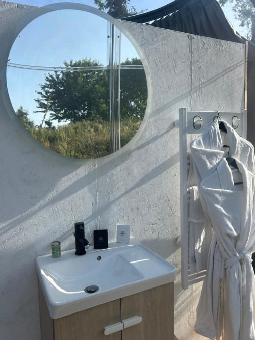 Salle de bains moderne de la chambre Topaze de l'hôtel Bubble Jade avec miroir circulaire, lavabo blanc, porte-serviettes et vue sur le jardin
