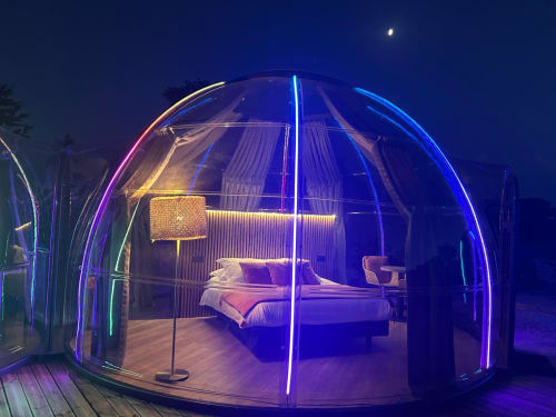La chambre en dôme géodésique illuminée de l'hôtel Bubble Jade la nuit, bordée de néon, avec lit et vue sur le jardin à Bazouges la Pérouse,