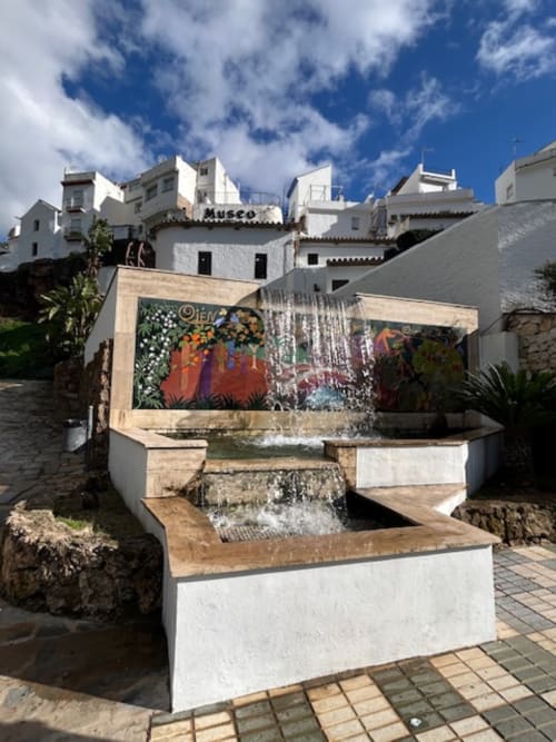 Fuente decorativa con azulejos coloridos en la plaza de Ojén, edificios encalados y señal de museo visible al fondo