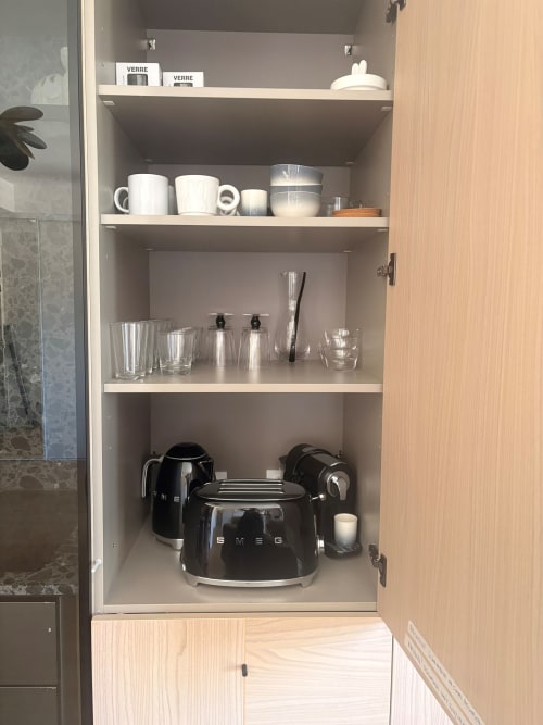 Hotel kitchen cabinet at Maisons Valmer Le Havre with organized shelves displaying white mugs, glasses, and black SMEG