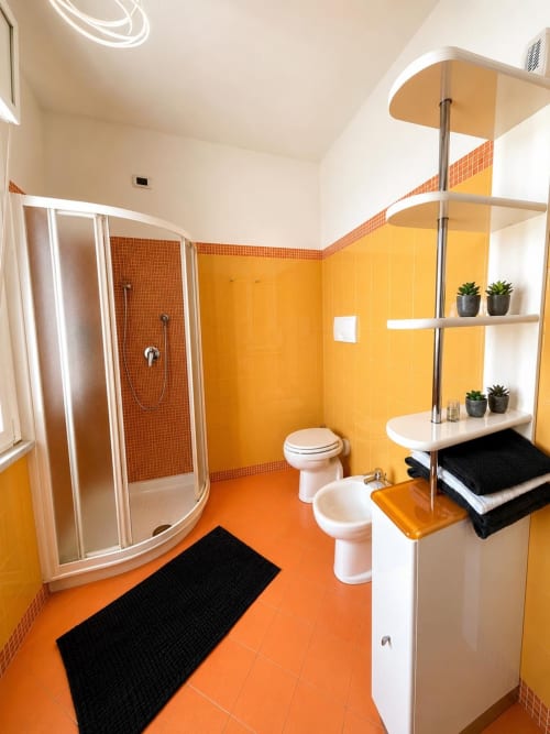 Bagno moderno a Bicocca Flat Milano con pareti arancioni vivaci, doccia piastrellata, bidet, WC e mensole con piante