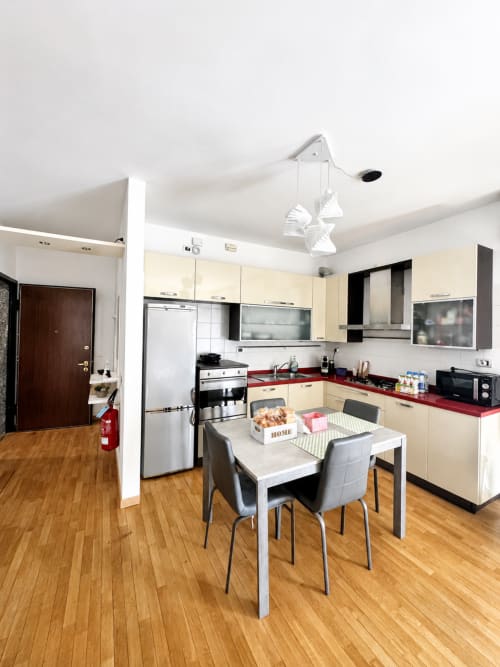 Moderna zona cucina-pranzo in Bicocca Flat B Milano con mobili color crema, piani di lavoro rossi, elettrodomestici in acciaio inox e dettagli in legno