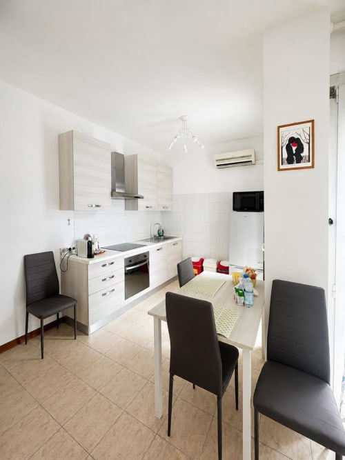 Cucina-soggiorno moderna nell'appartamento Bicocca Flat a Milano, con mobili bianchi, tavolo da pranzo, sedie nere e elettrodomestici