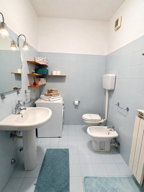 Moderno bagno con lavabo su colonna, bidet, wc, scaffali in legno, piastrelle azzurre e tappeto da bagno turchese a Bicocca Flat Milano