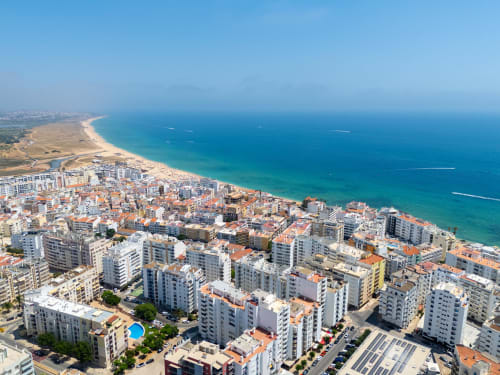Vista aérea da orla de Armação de Pêra com apartamentos coloridos, mar turquesa e praia arenosa sob céu azul