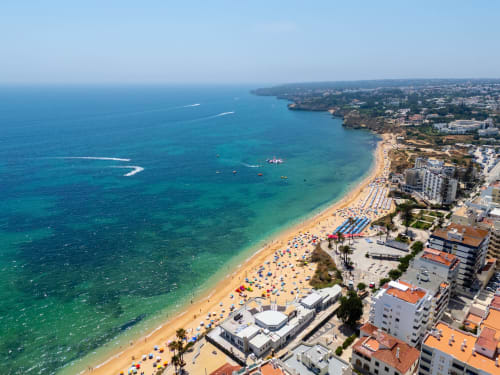 Vista aérea da praia de Armação de Pêra com areia dourada, águas azuis e edifícios brancos no litoral do Algarve