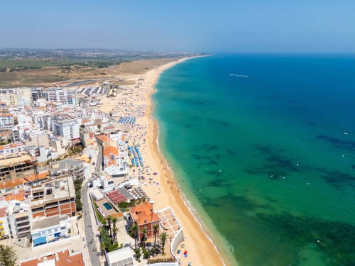 Vista aérea da praia de Armação de Pêra com águas turquesas, areia dourada e edifícios da vila costeira