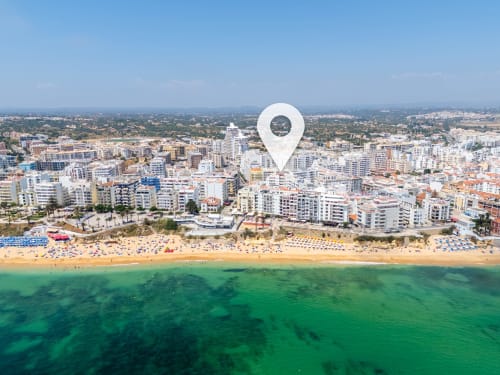 Vista aérea da praia de Armação de Pêra com águas turquesas, areia dourada e edifícios costeiros sob céu limpo