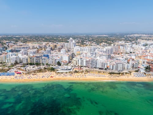 Vista aérea da Praia da Armação de Pêra com água turquesa, areia branca, guarda-sóis coloridos e edifícios brancos