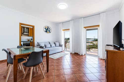 Sala luminosa com mesa de jantar, sofá, TV, cozinha e vista para varanda no Happy Nest AL, Armação de Pêra