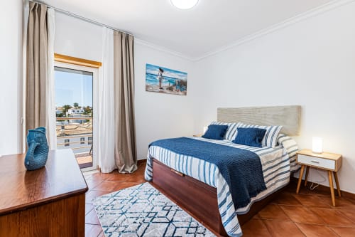 Quarto luminoso na Happy Nest AL em Armação de Pêra com roupa de cama azul, secretária de madeira, varanda com vista para o jardim e costa