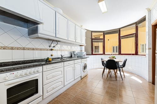 Cozinha moderna branca com bancadas pretas, mesa de jantar e grandes janelas no apartamento Marina Rocha em Portimão