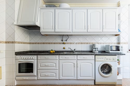 Cozinha moderna branca com fogão, máquina de lavar e bancada de granito escuro no apartamento Happy Nest AL em Portimão