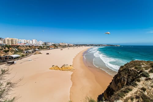 Praia de areia dourada com ondas turquesas em Portimão, Portugal, com falésias costeiras e apartamentos à beira-mar