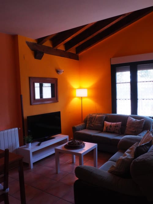 Luminoso salón naranja con sofá oscuro, mueble TV, lámpara de pie y vigas de madera vista en Senda del Duratón rural