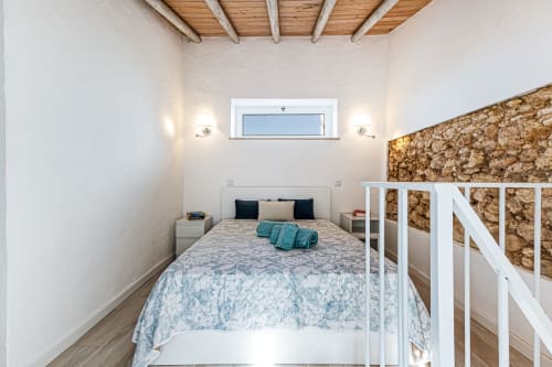 Quarto aconchegante da casa rural Casa Passarinho no Happy Nest AL, Pêra, com cama de ferro branca, vigas de madeira aparente e vista para o mar