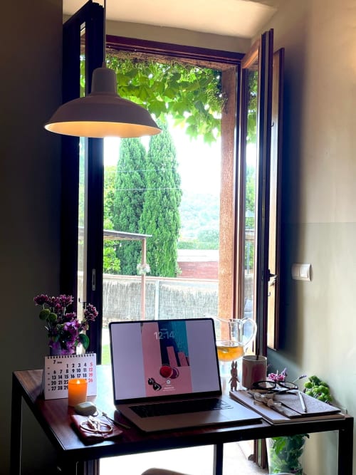 Espace de travail moderne dans un gîte de Costa Brava avec ordinateur portable, lampe suspendue, calendrier et vue sur le jardin par portes ouvertes