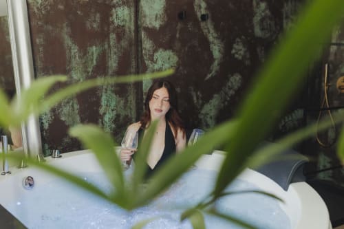 Donna nella vasca da bagno all'Oltre Exclusive Palermo, pianta verde in primo piano, sfondo con piastrelle rustiche