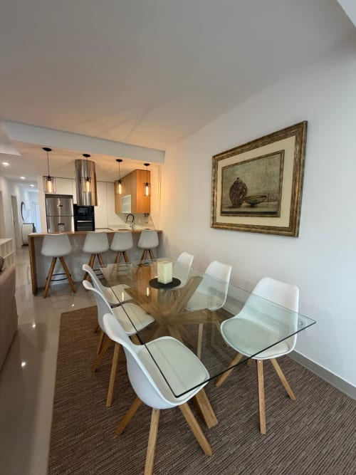 Área moderna de jantar e cozinha no apartamento Galagana Charm House com mesa de vidro, cadeiras brancas e iluminação suspensa