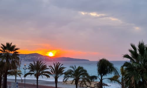 Sunset over Mediterranean Sea from Nice hotel balcony with palm trees and coastal mountains silhouetted against golden sky