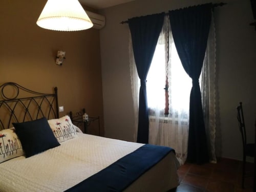 Acogedora habitación doble en Casa Rural El Infante en Oropesa con cama de hierro forjado, ropa de cama color crema, cortinas azul oscuro y ambiente cálido