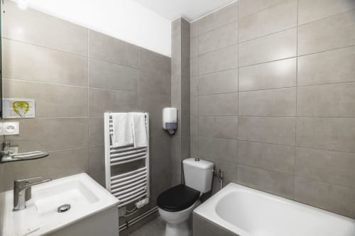 Salle de bain moderne aux murs en carrelage gris, lavabo blanc, toilettes à siège noir, sèche-serviettes chauffant et baignoire à l'Hôtel De La