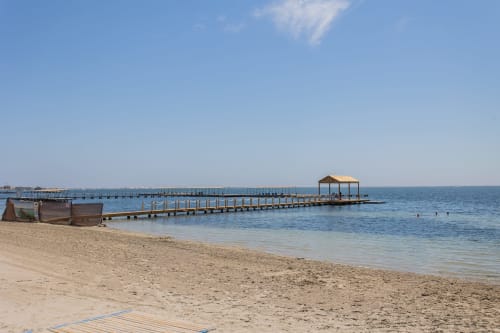 Muelle de madera que se extiende en el tranquilo mar azul de la playa San Javier con orilla arenosa y estructuras de embarcaciones