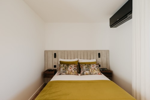 Quarto moderno em Villas Eden com roupa de cama amarela mostarda, almofadas estampadas, cabeceira bege e ar condicionado de parede