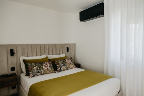 Quarto moderno em Villas Eden com cabeceira bege, roupa de cama branca, manta amarela e almofadas estampadas
