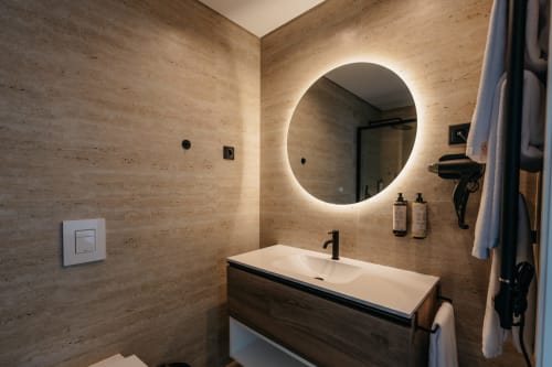 Banheiro moderno na Vila Rubi em Villas Eden com espelho redondo iluminado, paredes de mármore, vanity em madeira e acabamentos em preto.