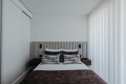 Quarto moderno na Villas Eden com cabeceira estofada cinza, roupa de cama escura, mesas de cabeceira em madeira e cortinas brancas translúcidas