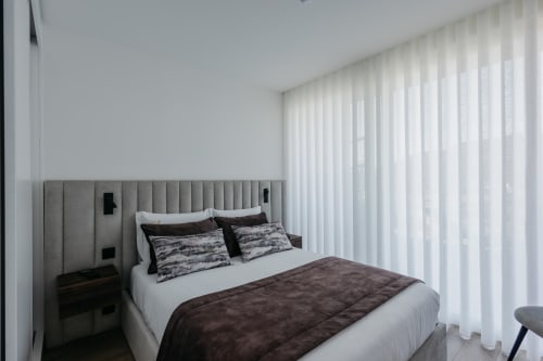 Quarto moderno em Villas Eden com cabeceira estofada cinza, roupa de cama escura e cortinas brancas translúcidas com vista portuguesa
