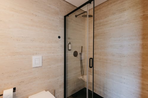 Banheiro moderno com divisória de vidro com moldura preta e paredes revestidas de travertino na Vila Quartz, Villas Eden