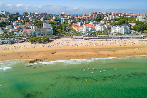 Vista de playa con arena, bañistas y olas, frente a coloridos edificios residenciales en una localidad costera española