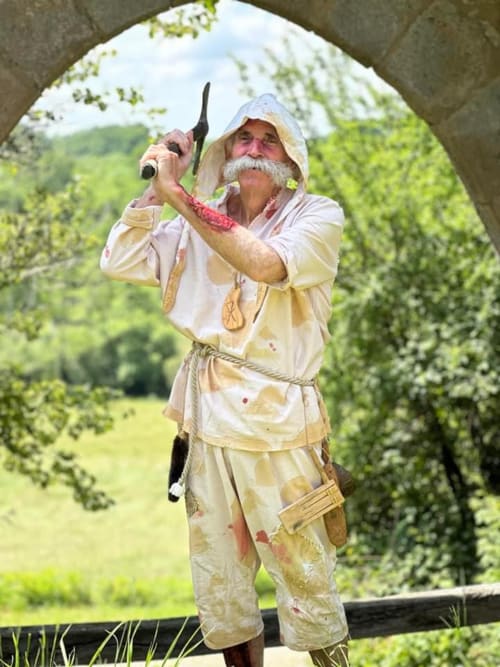 Homme barbu en costume d'époque tenant une canne à pêche sous une arche en pierre au Domaine de Châlus-Chabrol, Châlus