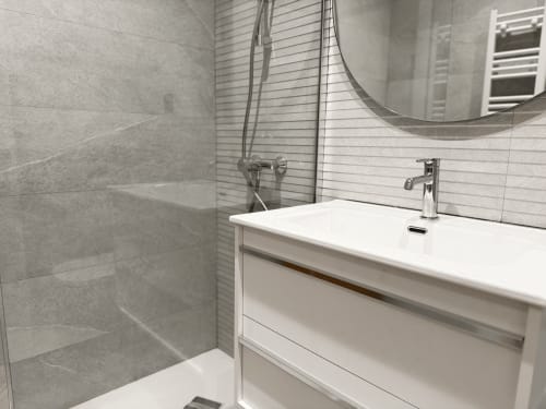 Baño moderno en el hotel MTM Rooms Santander con tocador blanco, espejo redondo, ducha abierta y paredes con azulejos grises